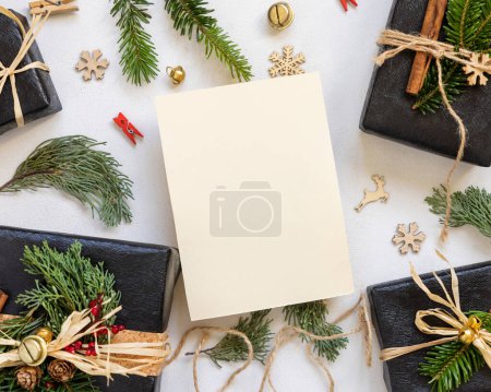 Téléchargez les photos : Carte en papier entre décor de Noël, coffrets cadeaux noirs et branches de sapin vert vue du dessus. Modélisation verticale de la carte blanche, pose à plat, espace de copie. Noël ou Nouvel An vœux de vacances - en image libre de droit