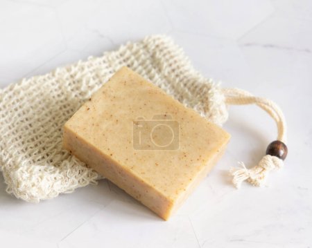 Photo for Beige handmade soap bar on sisal soap saver bag on white marble table close up, copy space. Natural herbal products for face and body care - Royalty Free Image
