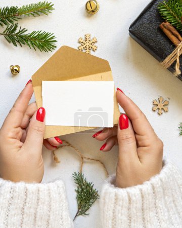 Photo for Hands with card and envelope near gift boxes, fir branches and Christmas decor, top view on white table. Winter composition with blank card, Mockup, copy space - Royalty Free Image