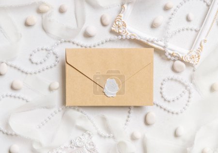 Photo for Sealed beige envelope near white frames, pearls, pebbles and silk ribbons on white table top view,  wedding mockup. Romantic scene with blank envelope flat lay. Valentines, Spring or Mothers day concept - Royalty Free Image