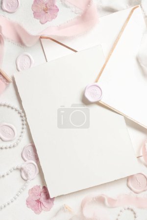 Photo for Card and envelope near pink decorations, seals and silk ribbons on white table top view,  wedding mockup. Romantic scene with vertical blank card flat lay. Valentines, Spring or Mothers day concept - Royalty Free Image