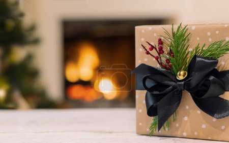 Téléchargez les photos : Coffret cadeau de Noël décoré avec des noeuds de ruban noir sur fond de cheminée en feu et arbre de Noël avec des lumières. Ambiance familiale d'hiver chaud, Backgroun vacances magiques - en image libre de droit