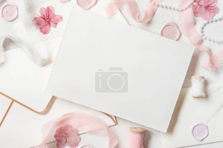 Photo for Paper card near pink decorations, seals and silk ribbons on white table top view,  wedding mockup. Romantic scene with horizontal blank card flat lay. Valentines, Spring or Mothers day concept - Royalty Free Image
