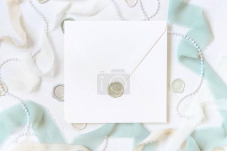 Photo for Sealed envelope near  light green and beige decorations and silk ribbons on white table top view,  Wedding mockup. Romantic scene with square blank envelope. Valentines, Spring or Mothers day stationery - Royalty Free Image