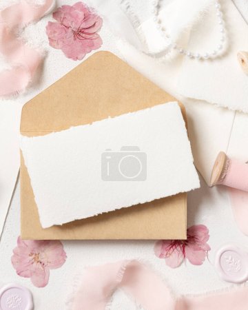 Photo for Card and envelope near pink decorations, seals and silk ribbons on white table top view,  wedding mockup. Romantic scene with horizontal blank card flat lay. Valentines, Spring or Mothers day concept - Royalty Free Image