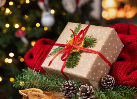 Téléchargez les photos : Cadeau de Noël avec arc près du pull rouge contre la cheminée en feu et les lumières du sapin de Noël. Ambiance familiale d'hiver chaude, Temps de vacances magique - en image libre de droit