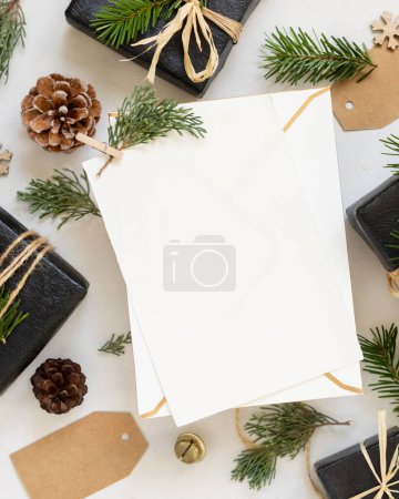 Téléchargez les photos : Carte en papier entre décor de Noël, coffrets cadeaux noirs et branches de sapin vert vue du dessus. Modélisation verticale de la carte blanche, pose à plat, espace de copie. Carte de voeux Noël ou Nouvel An - en image libre de droit