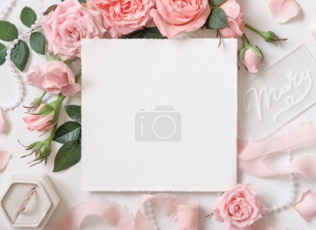Téléchargez les photos : Carte blanche près de roses rose clair, bague de fiançailles dans une boîte et rubans de soie sur table blanche vue sur le dessus, maquette de mariage. Couché plat romantique avec carte carrée et décor pastel - en image libre de droit