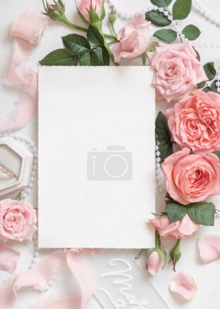 Foto de Tarjeta en blanco cerca de rosas rosadas claras, anillo de compromiso en una caja y cintas de seda en la vista superior de la mesa blanca, maqueta de boda. Romántica cama plana con tarjeta vertical y decoración pastel - Imagen libre de derechos