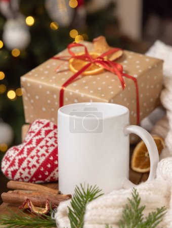 Photo for White coffee mug near Christmas present, cosy heart and sweater close up against Christmas tree lights and burning fireplace, copy space. Warm winter family atmospheric mock up, magical Holiday tim - Royalty Free Image