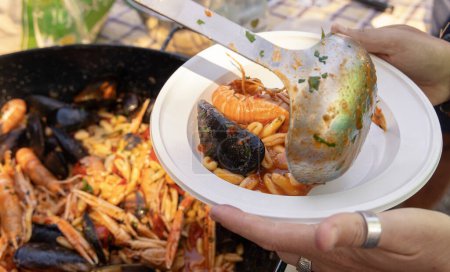 Hände halten einen Plastikteller Cavatelli mit schwarzen Miesmuscheln und Garnelen, während er mit der Schöpfkelle aus nächster Nähe serviert wird. Sommer-Familienpicknick mit italienischen Meeresfrüchten 