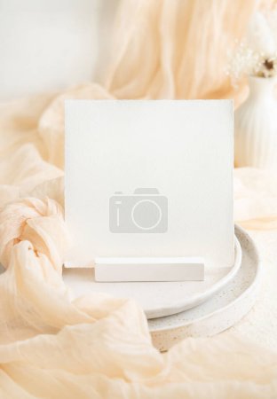 Photo for Square card near cream tulle fabric on plates on cream table close up. Wedding stationery mockup. Table decor with small vase and silk fabric knot, romantic interior - Royalty Free Image