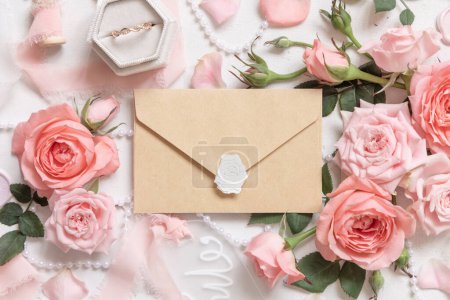 Photo for Kraft paper sealed envelope near light pink roses, engagement ring in a box and silk ribbons on white table top view,  wedding mockup. Romantic flat lay with pastel decor - Royalty Free Image