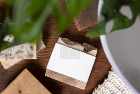 Photo for Beige handmade soap bar with blank label on bathroom wooden countertop near potted monstera plant and basin top view, packaging mockup, copy space. Natural herbal product for body hygiene - Royalty Free Image