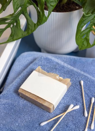 Photo for Handmade soap bar with blank label and bamboo cotton swabs on blue towels near basin and green monstera close up, mockup.  Lifestile scene with skincare hygiene product in bathroom - Royalty Free Image