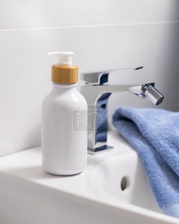Téléchargez les photos : Bouteille d'une pompe blanche avec robinet en bambou sur le lavabo près du robinet d'eau et serviette bleue pliée dans la salle de bain close up, maquette cosmétique. Composition minimale pour l'emballage de marque - en image libre de droit