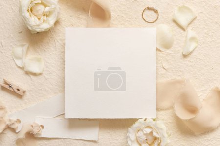 Quadratische Karte in der Nähe von cremefarbenen Rosen und Blütenblättern, Verlobungsring und Seidenbändern oben in beige, Attrappe. Romantisch flach mit Blanko-Karte und Pastell-Dekor 