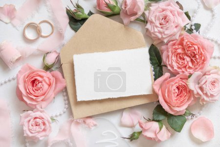 Foto de Tarjeta en blanco y sobre cerca de rosas de color rosa claro, anillos de boda y cintas de seda en la vista superior de la mesa blanca, papelería maqueta. Romántico piso con decoración pastel y flores - Imagen libre de derechos