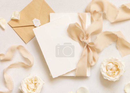 Photo for Cards tied with a beige silk ribbonnear cream roses and sealed envelope on white table top view, mockup. Romantic scene with vertical blank cards. Wedding, Valentines, Spring or Mothers day stationery - Royalty Free Image