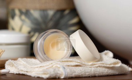 Téléchargez les photos : Pot de baume à lèvres ouvert avec couvercle vierge sur une serviette pliée à la crème près d'une plante en pot et d'un lavabo sur un comptoir de salle de bains en bois, emballage de marque cosmétique mocku - en image libre de droit