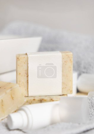 Téléchargez les photos : Barre de savon beige faite à la main avec étiquette vierge contre le lavabo, serviette et bouteilles de crème fermer, maquette. Scène de vie avec produits d'hygiène et de soins personnels dans la salle de bain - en image libre de droit