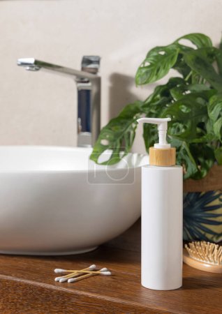 Photo for White one pump bottle near basin, green monstera, bamboo hair brush and cotton swabs on wooden countertop in bathroom, close up, cosmetic mockup. Modern scene with sustainable cosmetics or soaps - Royalty Free Image