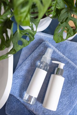 Photo for Cosmetic bottles on folded blue towels near basin and green monstera, top view, mockup.  Lifestile scene with skincare products in bathroom, Sustainable beauty routine - Royalty Free Image