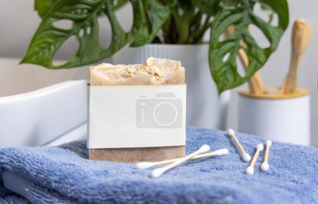 Téléchargez les photos : Savonnette artisanale avec étiquette vierge et coton-tige en bambou sur serviettes bleues près du lavabo et de la monstère verte en gros plan, maquette. Scène de vie avec des produits d'hygiène pour la peau dans la salle de bain - en image libre de droit