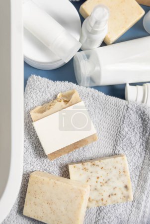 Téléchargez les photos : Porte-savon avec étiquette vierge sur serviette pliée grise près des bouteilles de crème et lavabo dans la salle de bain vue du dessus, maquette d'emballage, espace de copie. Produits d'hygiène naturels et cosmétiques - en image libre de droit
