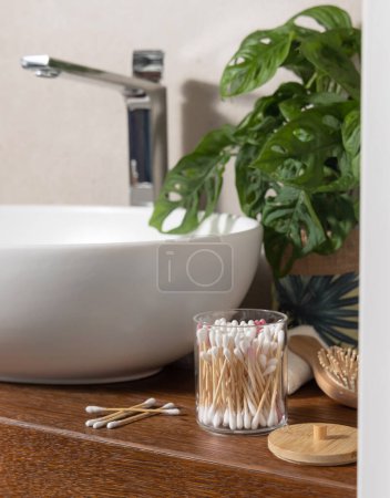 Photo for Bamboo cotton swabs in a jar near basin, green monstera, bamboo hair brush and cotton swabs on wooden countertop in bathroom, close up - Royalty Free Image