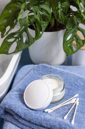 Photo for Opened cream jar with blank white lid and bamboo cotton swabs on blue towels near basin and green monstera blue countertop, close up, mockup.  Lifestile scene with skincare beauty product in bathroom - Royalty Free Image