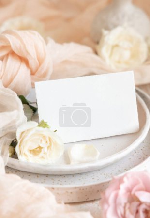 Téléchargez les photos : Petite carte horizontale près de tissu de tulle crème et roses sur des assiettes fermer. Modèle de papeterie de mariage. Carte de place de table vierge près de noeud en tissu de soie et fleurs, événement romantique - en image libre de droit
