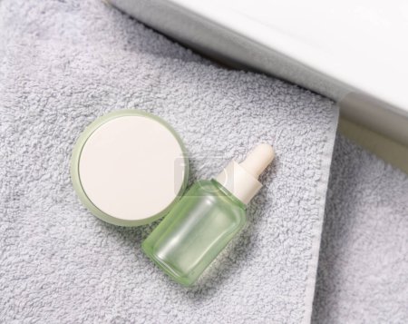 Translucent green dropper bottle and closed cream jar on light grey bath towel near basin top view, cosmetic mockup.  Beauty and skincare routines with natural organic products - serum and crea