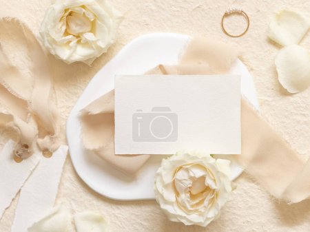Horizontale Karte in der Nähe von cremefarbenen Rosen und Blütenblättern, Verlobungsring und Seidenbändern von oben auf beige, Hochzeits-Attrappe. Romantische flache Liege mit leerer Papierkarte und Pastelldekor 