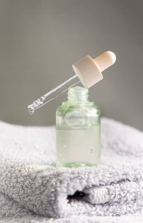 Photo for Drop falls from a pipette resting diagonally on a bottle filled with green liquid on grey folded towel in bathroom, close-up. Natural organic serum, oil or other cosmetic product for beauty routin - Royalty Free Image