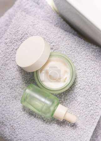 Translucent light green dropper bottle and opened cream jar on light grey bath towel near basin top view, cosmetic mockup.  Skincare routines with natural organic serum and crea