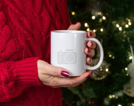 Photo for Woman in red sweater holding in hands white porcelain mug against Christmas tree with lights, mock up, copy space, front view. Girl with red nails drinking hot coffee or tea. Cozy holidays atmosphere - Royalty Free Image