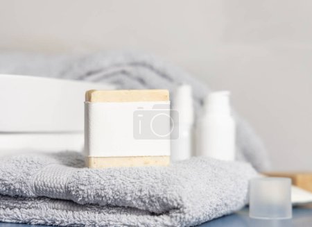 Téléchargez les photos : Barre de savon beige faite à la main avec étiquette vierge sur une serviette pliée gris clair contre le lavabo et les bouteilles cosmétiques fermer, maquette. Scène de vie avec produits d'hygiène et de soins personnels dans la salle de bain - en image libre de droit