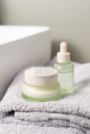 Translucent light green dropper bottle and closed cream jar on light grey folded bath towel near basin closeup, cosmetic mockup.  Beauty and skincare routines with natural organic products