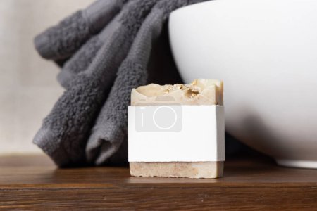 Jabonera con etiqueta en blanco cerca de toalla doblada gris en recipiente lavabo en encimera de madera marrón en baño beige, de cerca, maqueta para el embalaje de la marca. Producto natural de higiene artesanal
