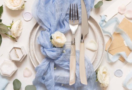 Foto de Mesa de boda con tenedor y cuchillo en placa, tela de tul azul claro, anillo de propuesta y flores de crema, vista superior. Decoración romántica de mesa pastel - Imagen libre de derechos