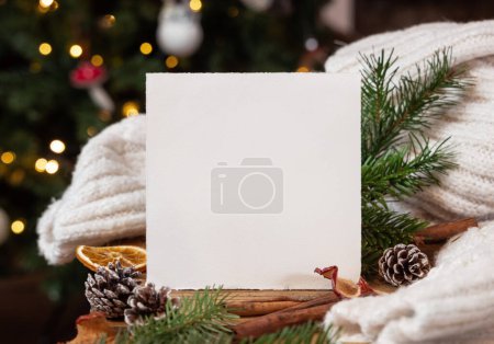 Tarjeta cuadrada cerca de la decoración de Navidad, acogedor suéter de punto blanco y ramas de abeto se cierran contra el centelleante árbol de Navidad, espacio para copiar. Cálido saludo de la familia de vacaciones, maqueta atmosférica