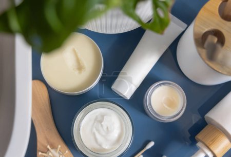 Photo for Opened cream jars and tubes on blue bathroom countertop near green plant and basin top view, packaging mockup, copy space. Natural personal care cosmetics and products - Royalty Free Image