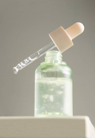 Photo for Drop falls from a pipette resting diagonally on a bottle filled with green liquid against gray backgound, close-up. Natural organic serum, oil or other cosmetic product for beauty routin - Royalty Free Image