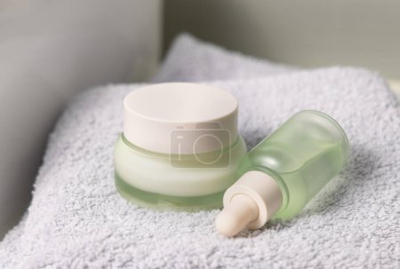 Translucent light green dropper bottle and closed cream jar on light grey folded bath towel near basin closeup, cosmetic mockup.  Beauty and skincare routines with natural organic products