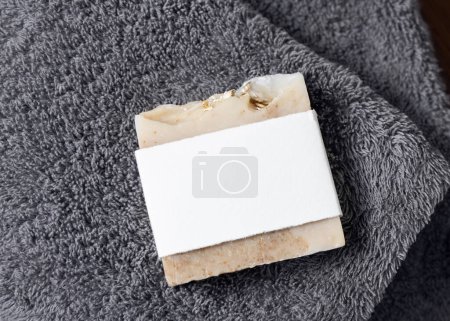 Photo for Beige handmade soap bar with blank label on dark grey folded towel top view, packaging mockup, copy space. Natural herbal hygiene products - Royalty Free Image