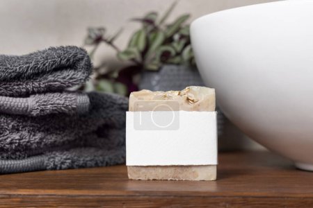Soap bar with blank label near grey folded towels, green plant and vessel basin on brown wooden countertop in beige bathroom, close up, brand packaging mockup. Natural handmade hygiene product