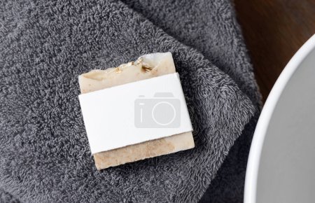Barra de jabón beige con etiqueta en blanco en toallas grises cerca del fregadero del recipiente en la encimera de madera marrón en el baño, vista superior, maqueta de embalaje de marca. Producto natural de higiene artesanal