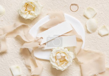 Small horizontal cards near cream roses and petals, engagement ring and silk ribbons top viewon beige,  mockup. Romantic flat lay with name cards and pastel decor 
