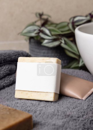 Barra de jabón con etiqueta en blanco en toallas plegadas de color gris oscuro, cerca de la planta verde y el recipiente en el baño, primer plano, maqueta de embalaje de la marca. Productos higiénicos